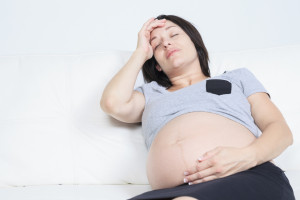Depressed pregnant woman holding hand on head and keeping eyes closed while sitting on a couch